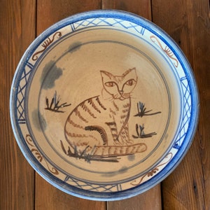 May include: A ceramic bowl with a hand-painted cat design. The bowl features a beige interior with a brown tabby cat illustration and blue accents. The rim has a blue and white geometric pattern and decorative elements.