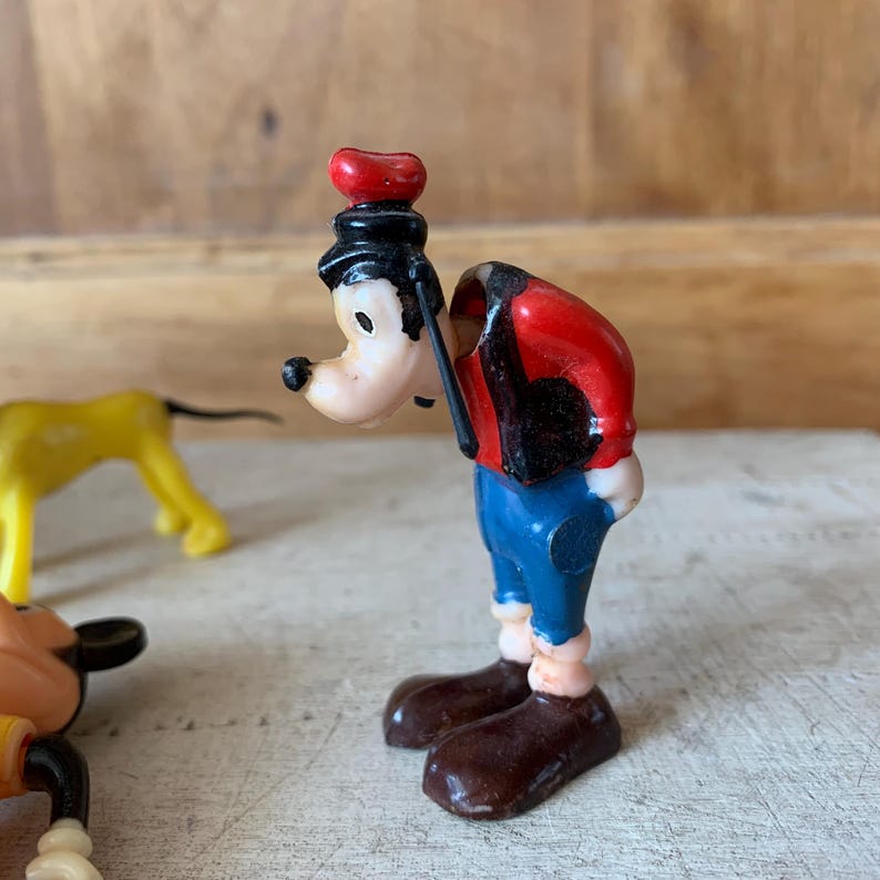 Puede incluir: Una figurita de pl&aacute;stico vintage de Goofy, un personaje de Disney, que lleva una camisa roja, pantalones azules y un sombrero negro. Est&aacute; de pie con la cabeza inclinada hacia abajo y los brazos a los lados.