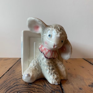 May include: A white ceramic bookend shaped like a lamb with pink ears and a pink and white checkered collar. The lamb is sitting on a white rectangular base.