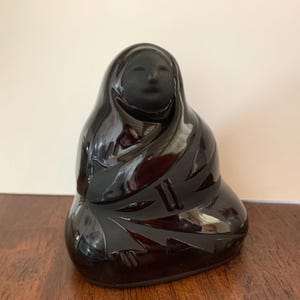 May include: A black ceramic sculpture of a seated figure with a draped head covering. The glossy finish reflects light, highlighting carved details on the figure's clothing. The sculpture is displayed on a wooden surface.