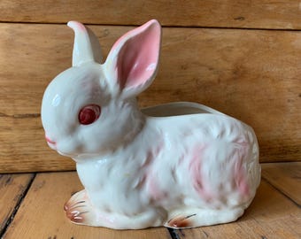 May include: White ceramic bunny planter with pink ears and red eyes. The bunny is sitting on its haunches with its paws visible.