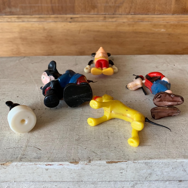 Puede incluir: Un conjunto de cinco figuras de personajes de Disney de pl&aacute;stico vintage. El conjunto incluye a Mickey Mouse, Pluto, Goofy y dos versiones de Mickey Mouse. Las figuras son de varios colores, incluyendo negro, rojo, amarillo, azul y marr&oacute;n.