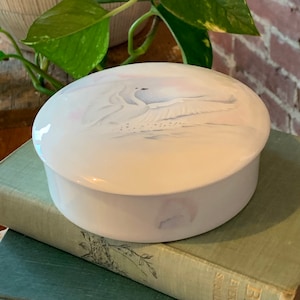 May include: White porcelain trinket box with a painted design of two white birds in flight. The box has a lid and is oval shaped.