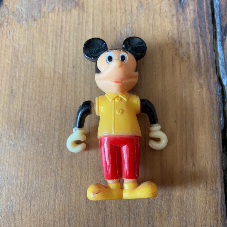 Puede incluir: Una figura vintage de pl&aacute;stico de Mickey Mouse con orejas negras, camisa amarilla, pantalones rojos y zapatos amarillos.