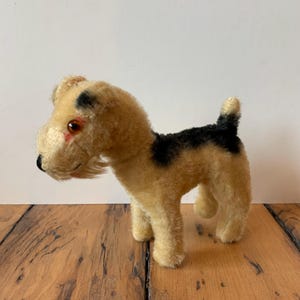 May include: A vintage stuffed dog toy with a tan body, black patches, and a black tail. The dog has a detailed face with brown eyes and a black nose. The toy is standing on a wooden surface.