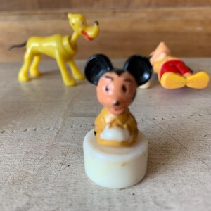 Puede incluir: Una figurita de pl&aacute;stico vintage de Mickey Mouse con una camisa amarilla y pantalones cortos negros. La figurita est&aacute; de pie sobre una base blanca.