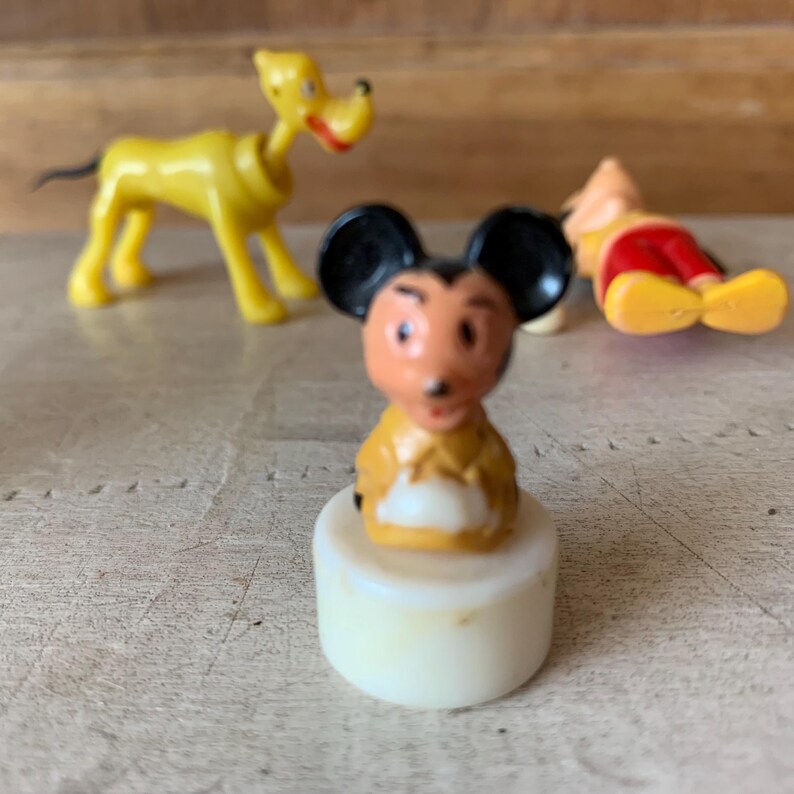 Puede incluir: Una figurita de pl&aacute;stico vintage de Mickey Mouse con una camisa amarilla y pantalones cortos negros. La figurita est&aacute; de pie sobre una base blanca.