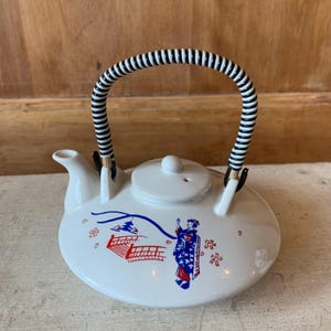 May include: White ceramic teapot with a black and white striped handle. The teapot has a floral design with a geisha and a red bridge.