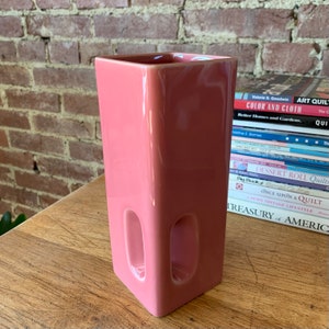 May include: A square, pink ceramic vase with two cut-out oval openings on the front. The vase is sitting on a wooden surface.