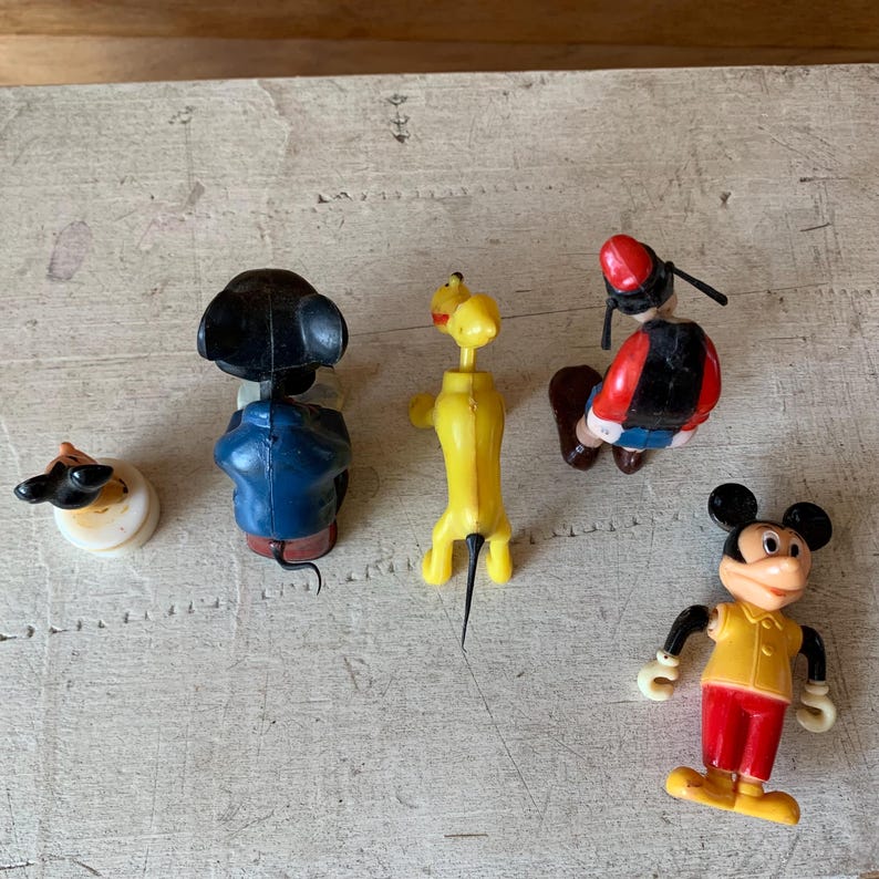 Puede incluir: Cinco figuritas de personajes de Disney de pl&aacute;stico vintage. Mickey Mouse lleva una camisa amarilla y pantalones rojos. Pluto es amarillo y tiene una cola negra. Una figurita de Mickey Mouse negra y azul est&aacute; tocando la bater&iacute;a. Una figurita de Mickey Mouse roja y negra lleva un sombrero rojo y sostiene un palo. Una figurita de Mickey Mouse negra y blanca lleva una camisa blanca y pantalones negros.