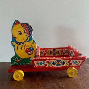 May include: A vintage wooden toy wagon with a cheerful yellow chick figure. The wagon is red with yellow wheels and floral painted details. The chick wears a bonnet and holds a basket with eggs. A charming Easter-themed collectible.