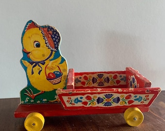 May include: A vintage wooden toy wagon with a cheerful yellow chick figure. The wagon is red with yellow wheels and floral painted details. The chick wears a bonnet and holds a basket with eggs. A charming Easter-themed collectible.