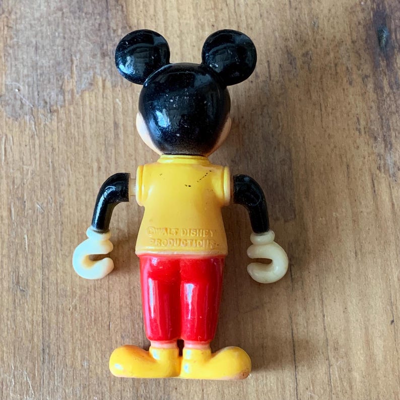 Puede incluir: Una figura vintage de pl&aacute;stico de Mickey Mouse con orejas negras, una camisa amarilla, pantalones rojos y guantes blancos. La figura tiene el texto "Walt Disney Productions" en la parte posterior.
