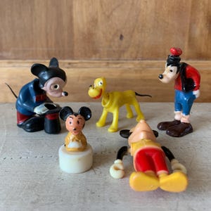 May include: A set of five vintage plastic Disney character figurines. The set includes Mickey Mouse, Pluto, Goofy, and two other Mickey Mouse figures. The figurines are all in good condition and have a classic look.