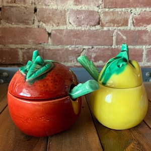 May include: A set of two ceramic sugar bowls shaped like a red tomato and a yellow pear. The bowls have green leaf lids and a matching green spoon.