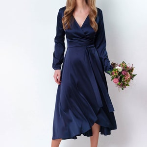 May include: A woman wearing a navy blue satin wrap dress with long sleeves and a tie at the waist. She is holding a bouquet of flowers.