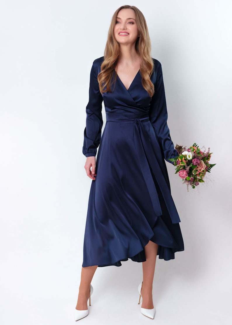 May include: A woman wearing a navy blue satin wrap dress with long sleeves and a tie at the waist. She is holding a bouquet of flowers.