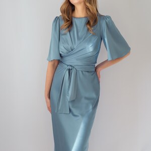 May include: A light blue satin dress with a wrap-style bodice and a tie at the waist. The dress features short, flared sleeves and a midi-length skirt. The model is smiling and has long, wavy hair.