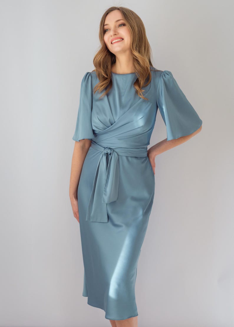 May include: A light blue satin dress with a wrap-style bodice and a tie at the waist. The dress features short, flared sleeves and a midi-length skirt. The model is smiling and has long, wavy hair.