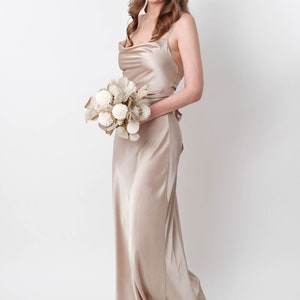 May include: A woman in a long, champagne-colored satin dress holds a bouquet of dried flowers. The dress has a cowl neck and thin straps.