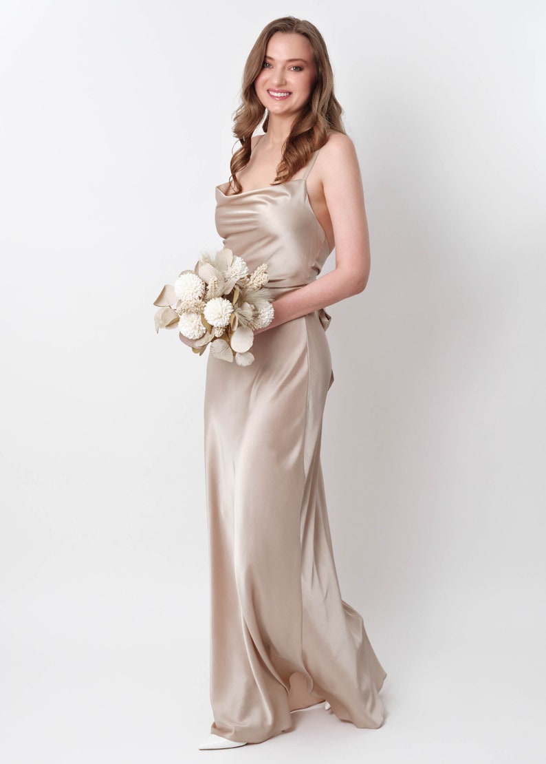 May include: A woman in a long, champagne-colored satin dress holds a bouquet of dried flowers. The dress has a cowl neck and thin straps.