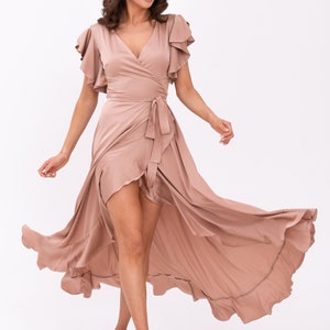 May include: A woman wearing a long, flowing, light brown wrap dress with ruffled sleeves. The dress has a high slit and a tie at the waist.