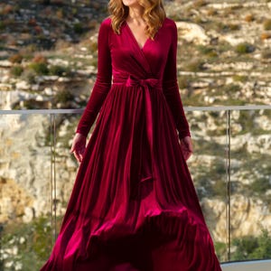 K&ouml;nnte beinhalten: Ein lang&auml;rmeliges, burgunderfarbenes Samtkleid mit Wickel-Oberteil und einer Schleife in der Taille. Das Kleid hat einen flie&szlig;enden Rock und wird von einer Frau getragen, die auf einem Balkon steht.
