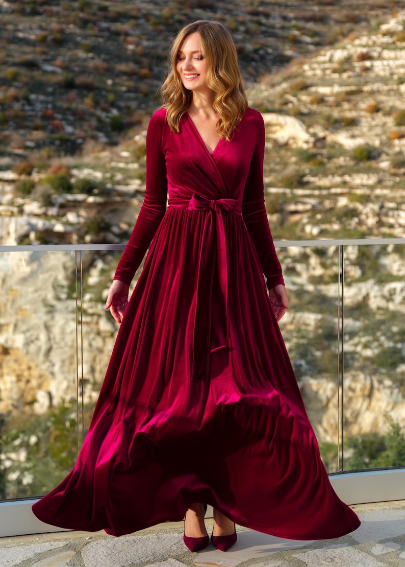 K&ouml;nnte beinhalten: Ein lang&auml;rmeliges, burgunderfarbenes Samtkleid mit Wickel-Oberteil und einer Schleife in der Taille. Das Kleid hat einen flie&szlig;enden Rock und wird von einer Frau getragen, die auf einem Balkon steht.