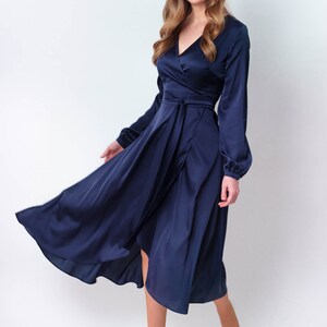 May include: A woman wearing a navy blue satin wrap dress with long sleeves. The dress has a V-neckline and a tie at the waist. She is wearing white heels.