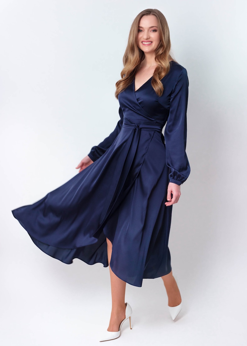 May include: A woman wearing a navy blue satin wrap dress with long sleeves. The dress has a V-neckline and a tie at the waist. She is wearing white heels.