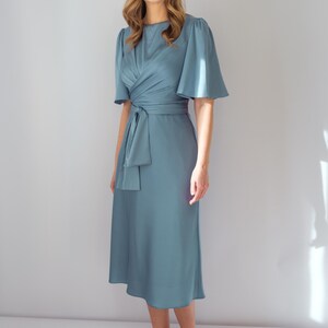 May include: A light blue midi dress with short, flared sleeves and a wrap-style bodice. The dress features a matching fabric belt tied at the waist and is paired with white heels. The dress is made of a smooth, flowing fabric.
