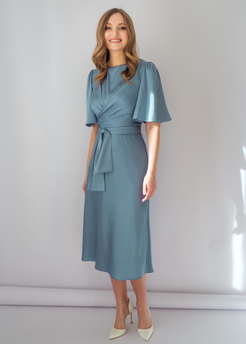 May include: A light blue midi dress with short, flared sleeves and a wrap-style bodice. The dress features a matching fabric belt tied at the waist and is paired with white heels. The dress is made of a smooth, flowing fabric.