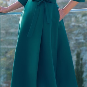 Puede incluir: Un vestido de color verde azulado con un corpi&ntilde;o cruzado y una falda fluida. El vestido tiene un cintur&oacute;n a juego en la cintura y mangas tres cuartos. El vestido tiene bolsillos y est&aacute; hecho de una tela lisa y s&oacute;lida.
