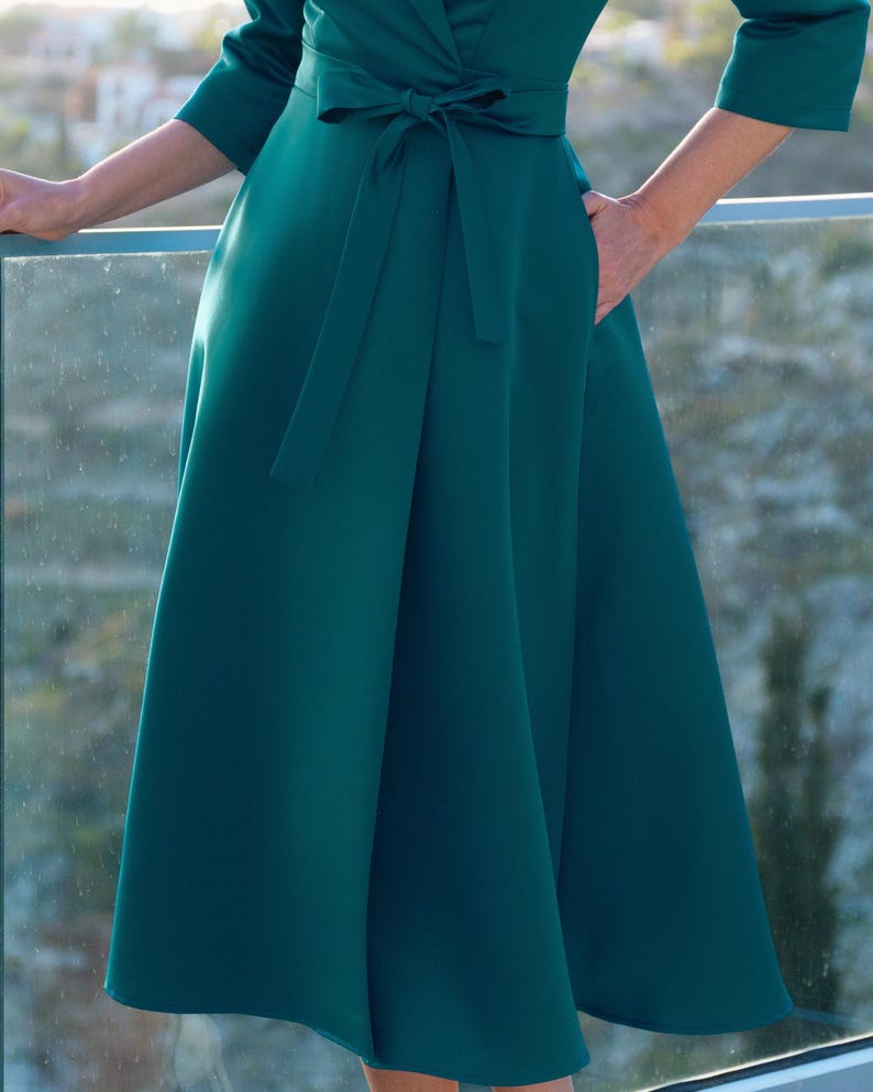 Puede incluir: Un vestido de color verde azulado con un corpi&ntilde;o cruzado y una falda fluida. El vestido tiene un cintur&oacute;n a juego en la cintura y mangas tres cuartos. El vestido tiene bolsillos y est&aacute; hecho de una tela lisa y s&oacute;lida.