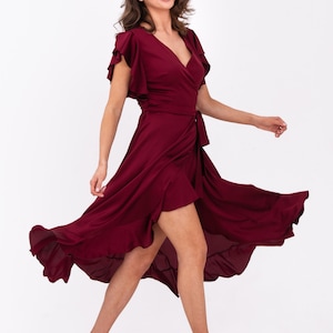 May include: A woman wearing a maroon wrap dress with ruffled sleeves and a high-low hemline. The dress has a tie at the waist and is flowing in the wind. She is wearing nude heels.