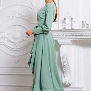 May include: A woman wearing a sage green wrap dress with a ruffled hem and long sleeves. She is standing in front of a white fireplace with ornate details.
