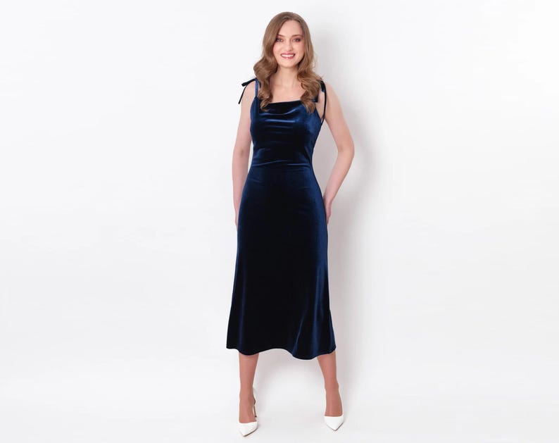 May include: A woman wearing a dark blue velvet midi dress with thin straps. The dress has a cowl neck and a fitted silhouette.