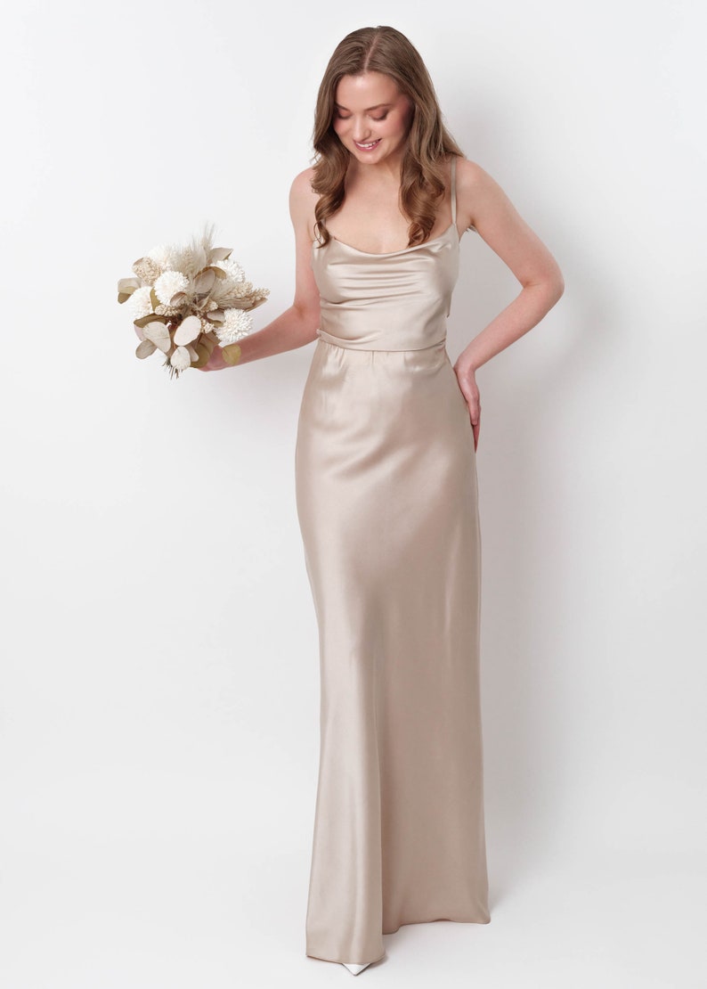 May include: A woman in a long, champagne-colored satin dress with a cowl neck and spaghetti straps. She is holding a bouquet of dried flowers.