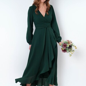 May include: A woman wearing a long, dark green wrap dress with long sleeves and a ruffled hem. She is holding a bouquet of flowers and smiling at the camera.
