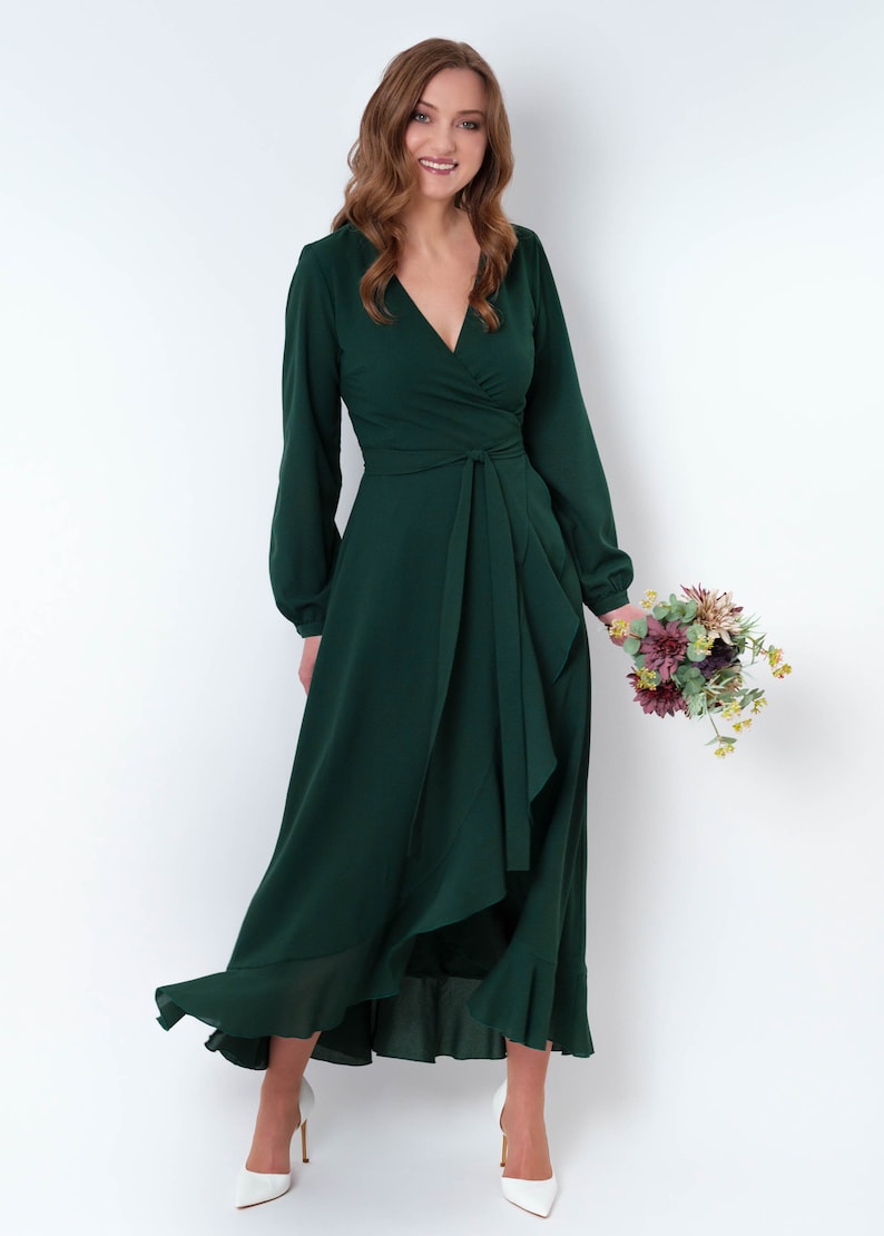May include: A woman wearing a long, dark green wrap dress with long sleeves and a ruffled hem. She is holding a bouquet of flowers and smiling at the camera.