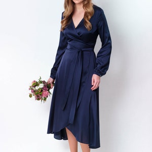 May include: A woman wearing a navy blue satin wrap dress with long sleeves and a tie at the waist. She is holding a bouquet of pink and white flowers.