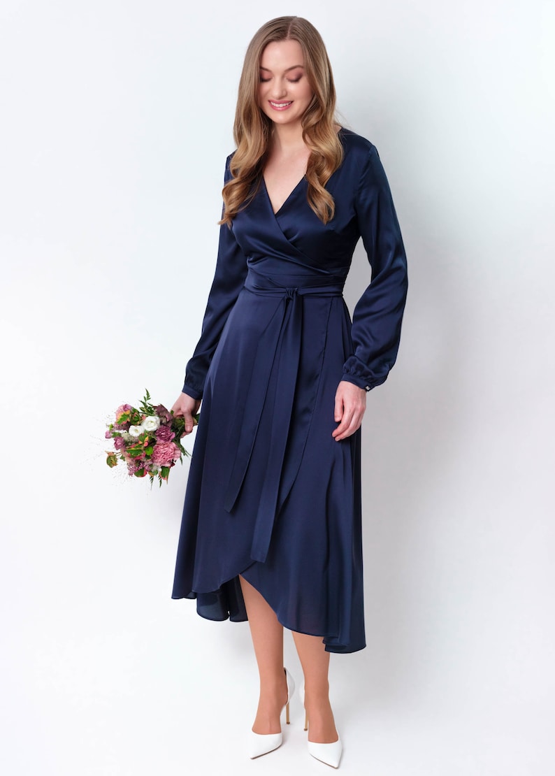 May include: A woman wearing a navy blue satin wrap dress with long sleeves and a tie at the waist. She is holding a bouquet of pink and white flowers.