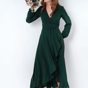 May include: A woman wearing a long, dark green wrap dress with a ruffled hem and long sleeves. She is holding a bouquet of flowers and is looking down at them.