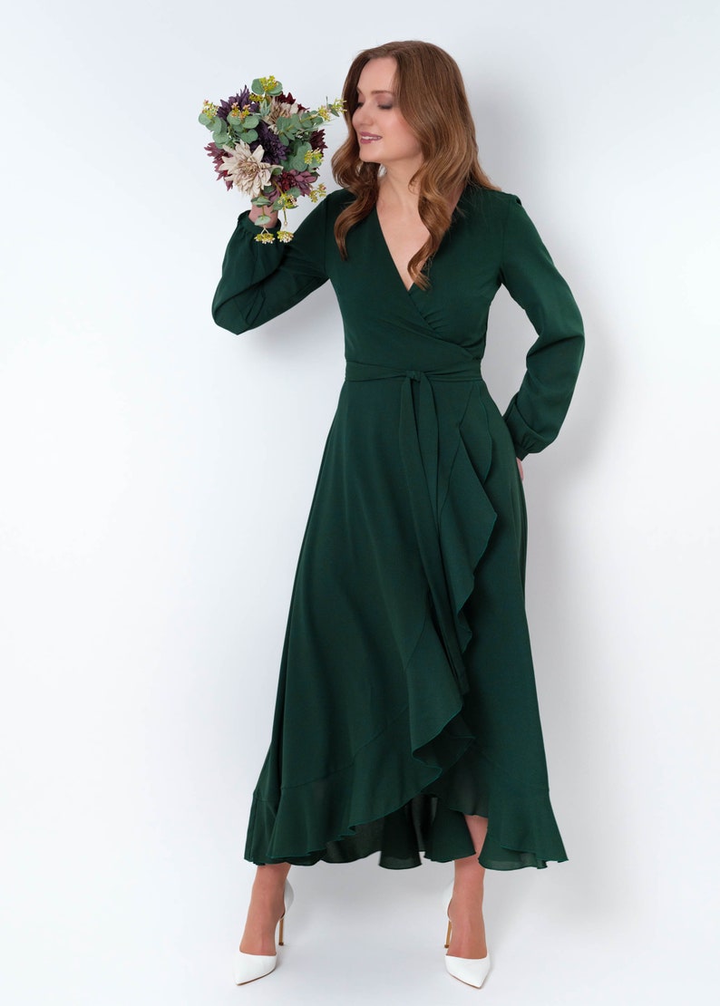 May include: A woman wearing a long, dark green wrap dress with a ruffled hem and long sleeves. She is holding a bouquet of flowers and is looking down at them.