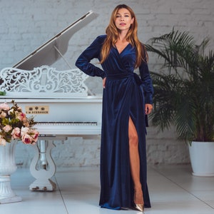 May include: A woman in a long, dark blue velvet dress with a side slit stands in front of a white grand piano. The piano has intricate carvings and the text "C.M. SCHROEDER" on the front. The woman is wearing gold heels and has long, wavy brown hair.