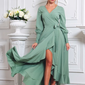 May include: A woman wearing a sage green wrap dress with long sleeves and a ruffled hem. The dress has a high slit on the left side and a tie belt at the waist. She is wearing silver heels.