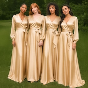 May include: Four women wearing long, flowing, champagne-colored dresses with long sleeves and wrap-style bodices. Each dress has a matching fabric belt tied at the waist. The women are standing in a grassy outdoor setting.