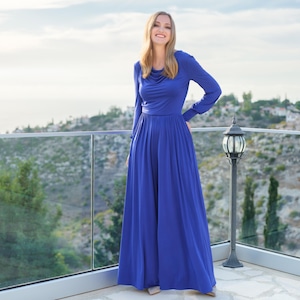 May include: A long, flowing cobalt blue dress with long sleeves and a cowl neckline. The dress is worn outdoors on a balcony overlooking a scenic landscape. The dress has a fitted bodice and a full, pleated skirt.