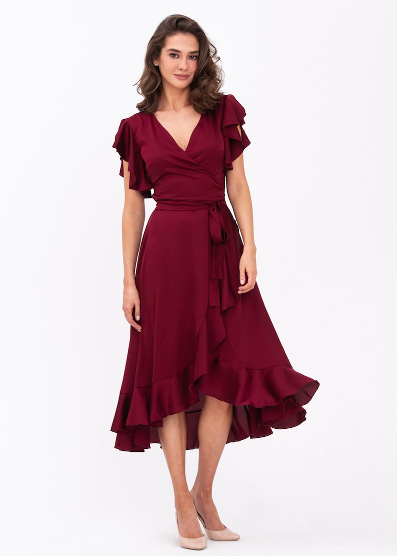 May include: A burgundy wrap dress with short ruffle sleeves and a high-low hemline. The dress has a tie at the waist and a ruffled skirt.