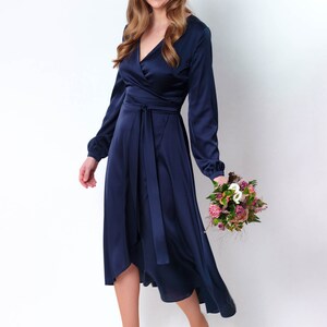 May include: A woman wearing a navy blue satin wrap dress with long sleeves and a high-low hemline. She is holding a bouquet of pink and white flowers.